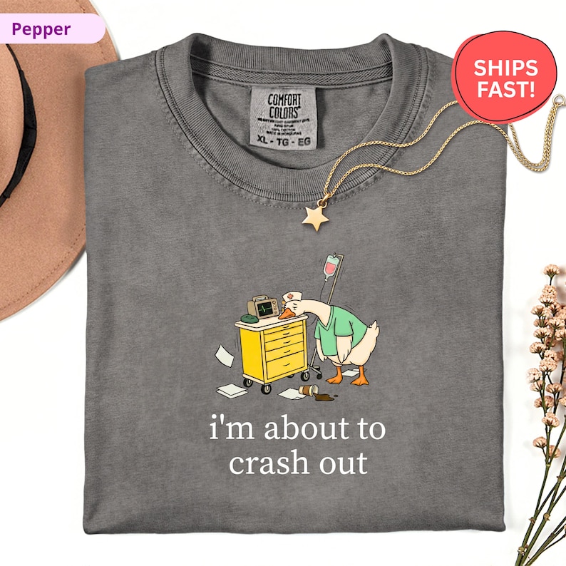 May include: A gray Comfort Colors t-shirt with a cartoon goose wearing a nurse's uniform, leaning on a medical cart. The text "i'm about to crash out" is below the graphic. A gold star necklace is on the shirt.