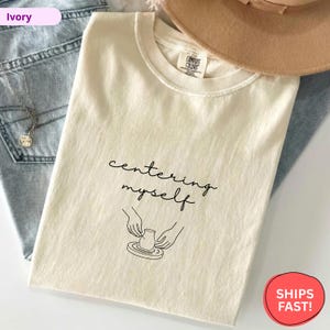 May include: Ivory-colored t-shirt with the phrase "centering myself" in a handwritten font, accompanied by a line drawing of hands shaping pottery. The shirt is displayed with blue jeans and a tan hat.