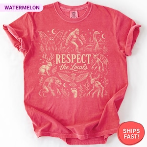 May include: A watermelon-colored short-sleeve t-shirt featuring a cream-colored graphic print. The design includes illustrations of cryptids and the text "RESPECT the Locals" in the center, surrounded by stars, moons, and forest elements.