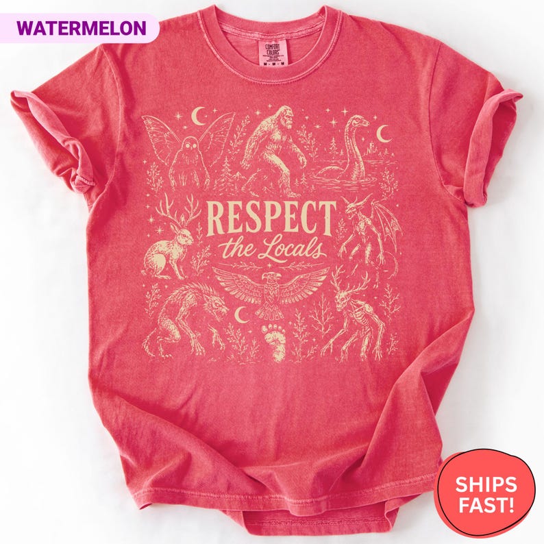 May include: A watermelon-colored short-sleeve t-shirt featuring a cream-colored graphic print. The design includes illustrations of cryptids and the text "RESPECT the Locals" in the center, surrounded by stars, moons, and forest elements.