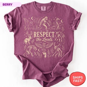 May include: A berry-colored short-sleeve t-shirt featuring a cream-colored graphic print with the text "RESPECT the Locals" surrounded by illustrations of mythical creatures including Bigfoot, a jackalope, a mothman, and a lake monster.