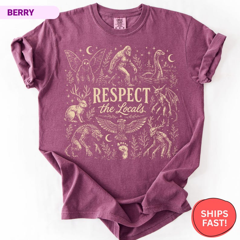 May include: A berry-colored short-sleeve t-shirt featuring a cream-colored graphic print with the text "RESPECT the Locals" surrounded by illustrations of mythical creatures including Bigfoot, a jackalope, a mothman, and a lake monster.