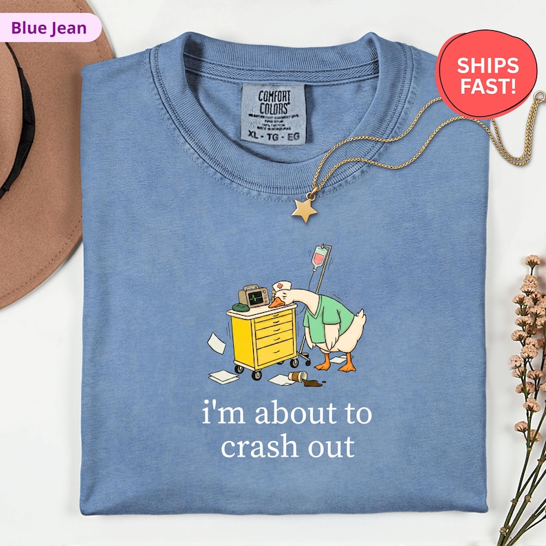 May include: Blue jean Comfort Colors t-shirt with a cartoon goose wearing a nurse's uniform, leaning on a medical cart. The text "i'm about to crash out" is printed below the graphic. A gold star pendant necklace is also visible.