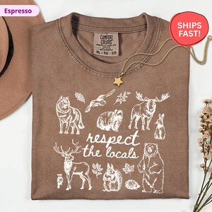May include: Espresso-colored t-shirt with a crew neck and the words "respect the locals" printed in white. The shirt features white illustrations of various animals, including a wolf, bear, and moose. A gold star pendant necklace is draped over the shirt.