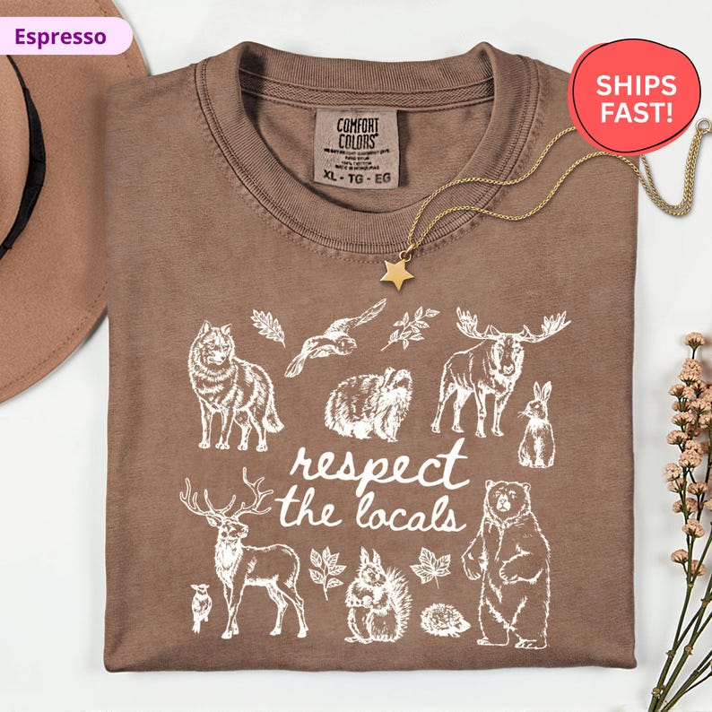 May include: Espresso-colored t-shirt with a crew neck and the words "respect the locals" printed in white. The shirt features white illustrations of various animals, including a wolf, bear, and moose. A gold star pendant necklace is draped over the shirt.