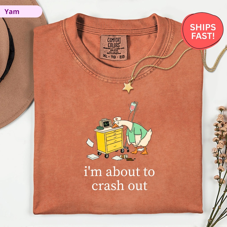 May include: Rust-colored t-shirt with a cartoon goose wearing a nurse's uniform, leaning on a yellow medical cart. The text "i'm about to crash out" is printed below the graphic. A gold star pendant hangs from the neckline.