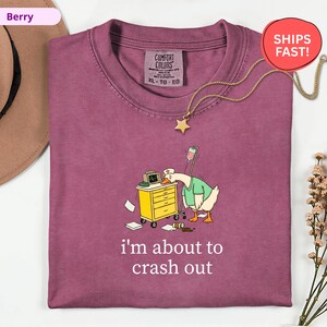 May include: Berry-colored Comfort Colors t-shirt with a cartoon goose wearing a nurse's uniform, leaning against a medical cart. The text "i'm about to crash out" is printed below the goose. A gold star pendant necklace is also visible.