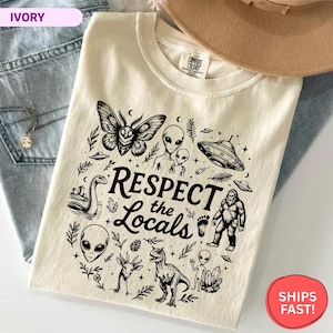 May include: An ivory t-shirt featuring a black graphic print with the text "RESPECT the Locals" surrounded by illustrations of a moth, aliens, a UFO, the Loch Ness monster, Bigfoot, a dinosaur, crystals, and various botanical elements.