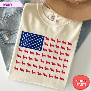 Puede incluir: Camiseta de color marfil con un diseño de la bandera estadounidense. El campo azul tiene huellas de patas blancas, y las rayas están compuestas por siluetas de perros corgi rojos. Un sombrero beige y unos vaqueros azules están en el fondo.