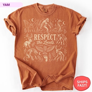 May include: A rust-colored short-sleeve t-shirt featuring a cream-colored graphic print of various mythical creatures, including Bigfoot and the Loch Ness Monster, with the text "RESPECT the Locals" centered among the illustrations.