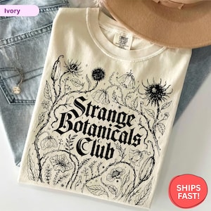 以下が含まれることがあります： 黒い花柄と「Strange Botanicals Club」の文字が入ったアイボリー色のスウェットシャツ。デザインは様々な植物のイラストが特徴です。スウェットシャツはブルージーンズの上に折りたたまれています。