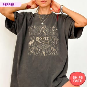 May include: A charcoal gray short-sleeve t-shirt featuring a beige graphic print of various cryptids and forest creatures. The central text reads "RESPECT the Locals" surrounded by illustrations of Bigfoot, a jackalope, and a mothman.