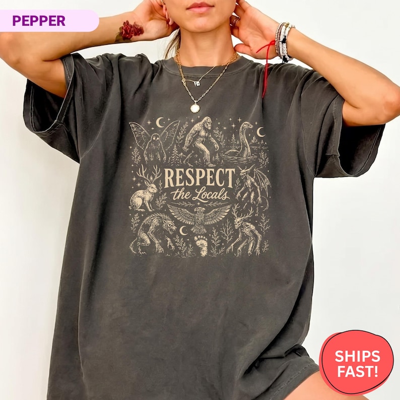 May include: A charcoal gray short-sleeve t-shirt featuring a beige graphic print of various cryptids and forest creatures. The central text reads "RESPECT the Locals" surrounded by illustrations of Bigfoot, a jackalope, and a mothman.