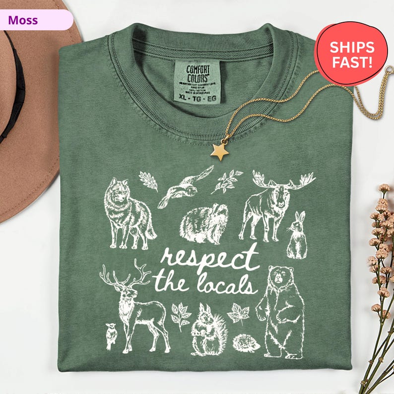 May include: A moss green t-shirt with a white graphic of various forest animals and the text "respect the locals." The shirt has a crew neck and short sleeves. A gold star pendant necklace is draped on the shirt.