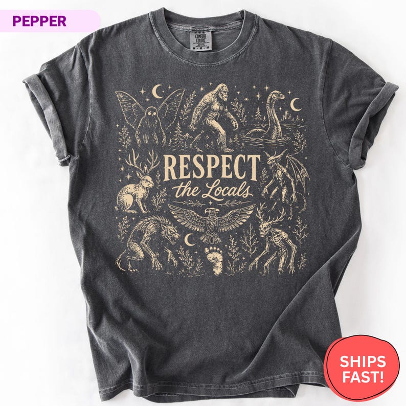 May include: A charcoal gray short-sleeve t-shirt featuring a cream-colored graphic print of various cryptids and mythical creatures. The central text reads "RESPECT the Locals" surrounded by illustrations of Bigfoot, a jackalope, and a mothman.