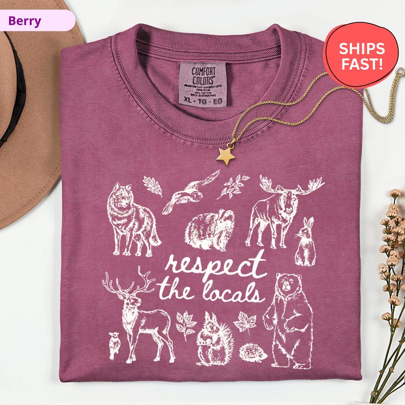 May include: Berry-colored t-shirt with a white graphic of various animals and the text "respect the locals." The shirt has a crew neck and short sleeves. A gold star pendant necklace is draped over the shirt.