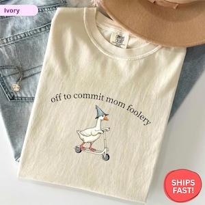 May include: Ivory-colored t-shirt with the phrase "off to commit mom foolery" above a cartoon goose riding a scooter and wearing a wizard hat. The shirt is made of a soft material.