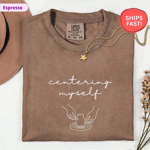 May include: Espresso-colored t-shirt with the words "centering myself" in white script. The shirt features a graphic of hands shaping a pot on a pottery wheel. A gold star pendant necklace rests on the shirt.