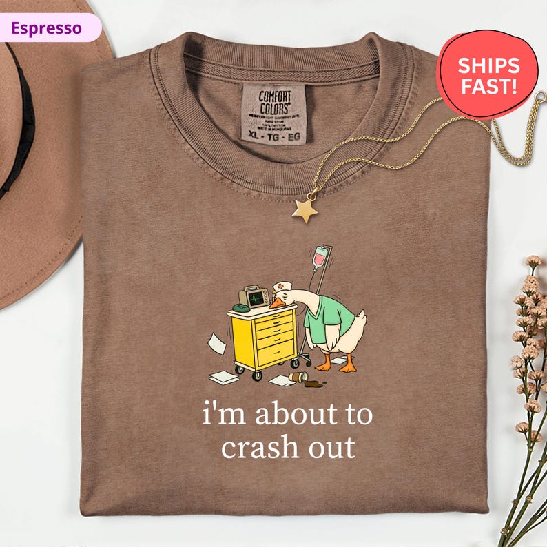 May include: Espresso-colored t-shirt with a cartoon goose wearing a nurse's uniform, leaning on a medical cart. The text "i'm about to crash out" is printed below the graphic. A gold star pendant necklace is draped on the shirt.