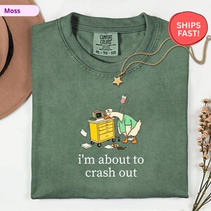 May include: A moss green Comfort Colors t-shirt with a cartoon goose wearing a nurse's uniform, leaning on a medical cart. The text "I'm about to crash out" is printed below the graphic. A gold star pendant hangs from the neckline.