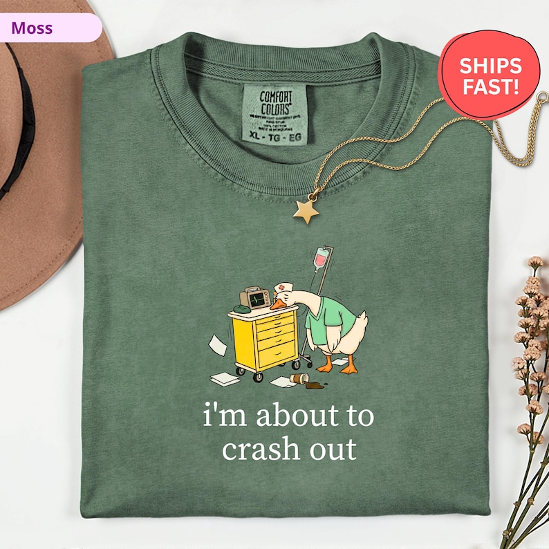 May include: A moss green Comfort Colors t-shirt with a cartoon goose wearing a nurse's uniform, leaning on a medical cart. The text "I'm about to crash out" is printed below the graphic. A gold star pendant hangs from the neckline.