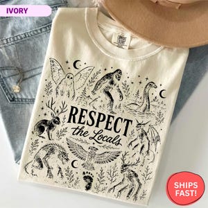 May include: An ivory t-shirt featuring black ink illustrations of mythical creatures including Bigfoot, a jackalope, a mothman, and a dragon. The text "RESPECT the Locals" is printed in the center. The shirt has a crew neckline and a Comfort Colors label.