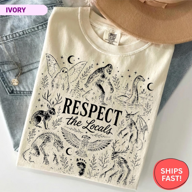 May include: An ivory t-shirt featuring black ink illustrations of mythical creatures including Bigfoot, a jackalope, a mothman, and a dragon. The text "RESPECT the Locals" is printed in the center. The shirt has a crew neckline and a Comfort Colors label.