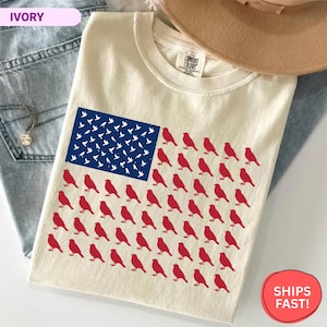 May include: An ivory t-shirt featuring a graphic of the American flag composed of small red bird silhouettes in the stripes and white bird silhouettes in the blue field. The shirt has a crew neckline and a Comfort Colors brand label at the neck.