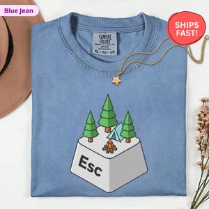 May include: A blue jean Comfort Colors t-shirt with a graphic of a computer 'Esc' key with a camping scene. The scene includes green trees, a blue tent, and a campfire. A gold star necklace is draped over the shirt.