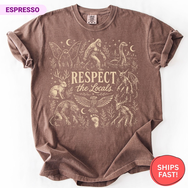 May include: An espresso-colored short-sleeve t-shirt featuring a cream-colored graphic print of various cryptids and mythical creatures. The central text reads "RESPECT the Locals" surrounded by illustrations of Bigfoot, a jackalope, and a mothman.