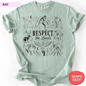 May include: A light green t-shirt featuring a black graphic print of various mythical creatures including Bigfoot, a jackalope, and a dragon. The text "RESPECT the Locals" is centered in the design, surrounded by stars, moons, and trees.
