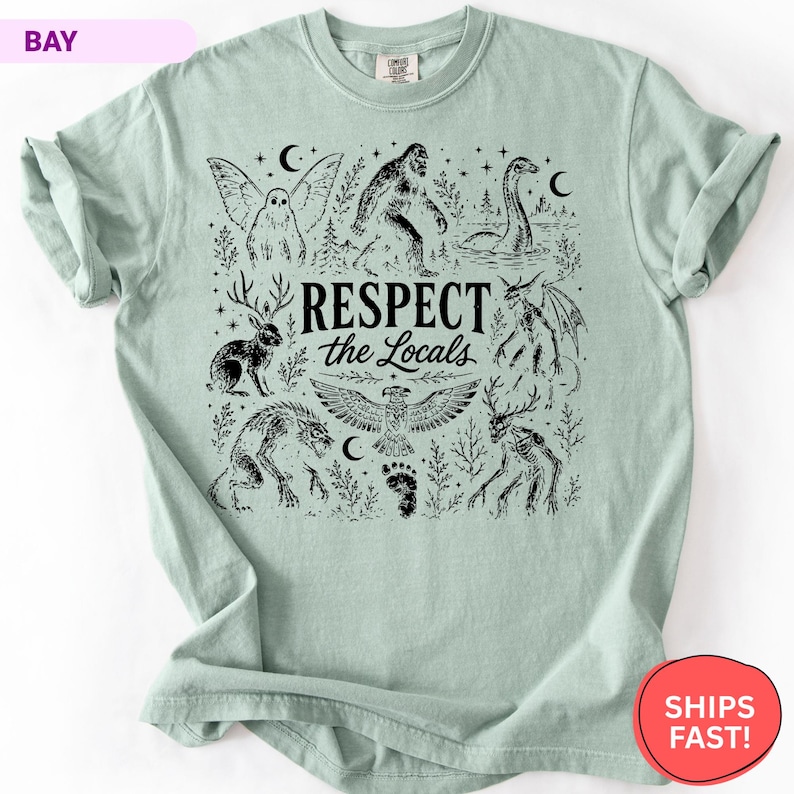 May include: A light green t-shirt featuring a black graphic print of various mythical creatures including Bigfoot, a jackalope, and a dragon. The text "RESPECT the Locals" is centered in the design, surrounded by stars, moons, and trees.
