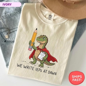 Könnte beinhalten: Elfenbeinfarbenes T-Shirt mit einem Cartoon-Dinosaurier, der einen Bleistift hält und einen roten Umhang trägt. Der Dinosaurier hat einen Klemmbrett mit der Aufschrift "School Support IEP Notes". Der Text "We Write IEPs at Dawn" ist darunter gedruckt.