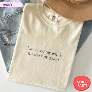May include: Ivory-colored t-shirt with the text "i survived my wife's master's program" in black. The shirt is made of a soft material and is folded on top of a pair of blue jeans and a tan hat.