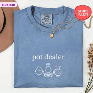 May include: A blue jean Comfort Colors t-shirt with the text "pot dealer" and a graphic of three pottery pieces. A gold star pendant necklace is draped across the shirt. The shirt is folded and has a tag that says XL-TG-EG.