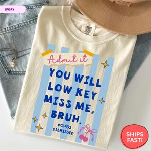 May include: Ivory-colored t-shirt with the text "Admit it, YOU WILL LOW KEY MISS ME, BRUH." in blue and pink. The shirt features blue vertical stripes, yellow stars, and a cherry graphic. The shirt is folded on top of blue jeans.