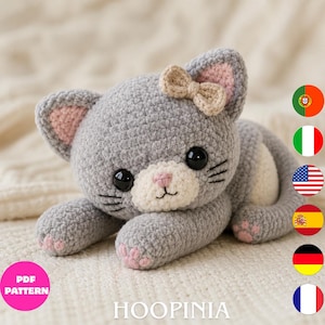 May include: A gray crocheted cat plush toy with a white muzzle, pink inner ears, and pink paw pads. The cat features large black eyes, black embroidered whiskers, and a small beige bow on its head. The text PDF PATTERN and HOOPINIA are visible.