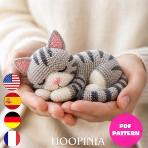 Peut inclure: Un petit chat endormi au crochet, réalisé avec du fil gris et blanc à rayures gris foncé. Le chat présente des oreilles intérieures roses, un nez rose et des yeux fermés. Le texte HOOPINIA et PDF PATTERN est visible sur l'image.
