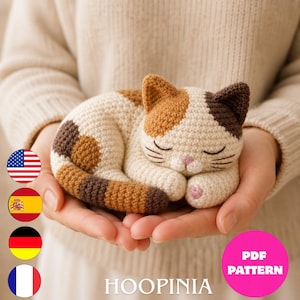 Peut inclure: Une petite figurine de chat calico crochetée avec des taches blanches, fauves et brun foncé. Le chat est recroquevillé en position de sommeil avec les yeux fermés, des moustaches brodées et un petit nez rose. Le texte HOOPINIA et PDF PATTERN est visible.