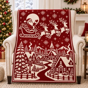 May include: A deep red and white knit throw blanket featuring a festive holiday scene. The design displays Santa Claus in a sleigh pulled by three reindeer above a snowy village with houses, a church, pine trees, and falling snowflakes.