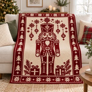 Puede incluir: Una manta de punto color crema y rojo oscuro con una figura central de cascanueces, rodeada de adornos colgantes, copos de nieve y cajas de regalo, con un borde de diseño repetitivo de abetos y copos de nieve.