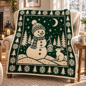 Puede incluir: Una manta de punto verde oscuro y crema con un muñeco de nieve central que lleva gorro y bufanda, sosteniendo un farol. El diseño incluye un conejo, pinos, una luna creciente y copos de nieve, con un borde decorativo de árboles y estrellas.