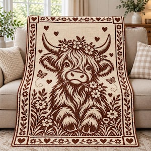 Puede incluir: Una manta rectangular con un diseño en color marrón oscuro y crema. El motivo muestra una vaca de las Tierras Altas central con una corona de flores, rodeada de pequeños corazones, mariposas y flores, todo dentro de un borde decorativo.