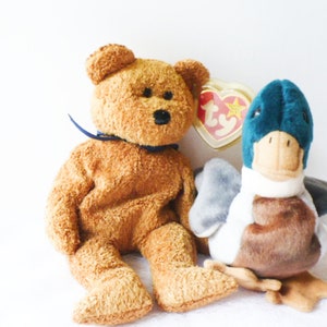 May include: A brown teddy bear with a blue ribbon around its neck and a teal blue duck with a brown beak and wings. Both are plush toys with Ty tags.