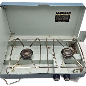 Può includere: Fornello da campeggio Primus vintage, aperto, con esterno azzurro. Il fornello presenta due bruciatori, griglie metalliche e manopole di controllo. Il logo Primus e le informazioni sul prodotto sono visibili all'interno del coperchio.