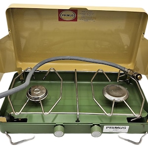 May include: A green and beige Primus 2397 camping stove with two burners. The stove has a hinged lid and a gray gas line. The Primus logo is visible on the lid and the front of the stove.