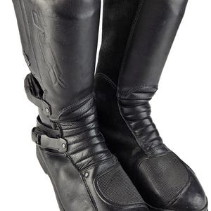 Könnte beinhalten: Ein Paar schwarze Leder-Motorrad-Stiefel mit einem Reißverschluss an der Seite und einem Schnallenriemen. Die Stiefel haben ein strukturiertes Design und eine dicke Sohle.