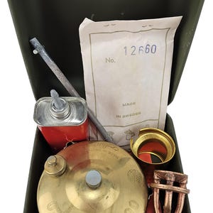 Könnte beinhalten: Eine grüne Metallbox mit einem Messing-Optimus No. 00 Kocher, einem roten Metall-Kraftstoffflasche, einer Metallstange, einem Messingbrenner und einer Messingkappe. Die Box hat ein Papieretikett mit dem Text "No. 12660 Hergestellt in Schweden".