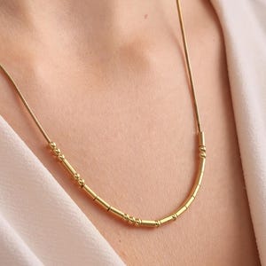 May include: A gold-colored necklace with a delicate chain and a curved bar pendant. The pendant features small, decorative beads. The necklace is worn against a cream-colored blouse.