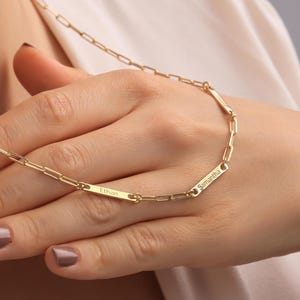 May include: A gold-colored chain necklace with two rectangular bars engraved with the names "Ethan" and "Samantha." The necklace is a delicate design with a link chain. The necklace is being worn.